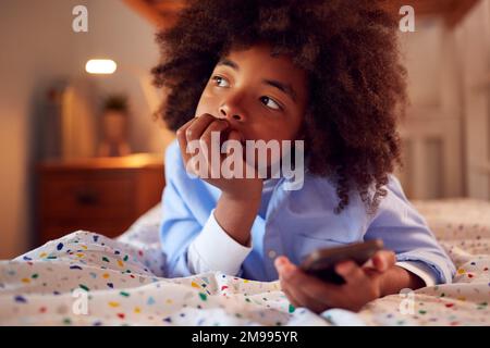 Worried Boy In Bedroom Lying On Bed Holding Mobile Phone Being Bullied Online Stock Photo