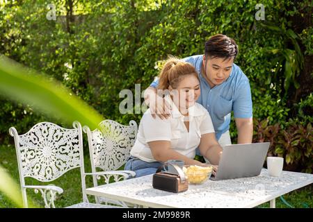 Young Asian chubby couple using a laptop in the outdoors green park together. Happy smiling man and woman working remotely and starting own business together Stock Photo