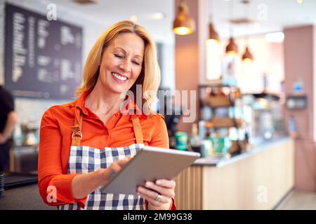 Mature Female Owner Of Coffee Shop Or Restaurant Using Digital Tablet Stock Photo