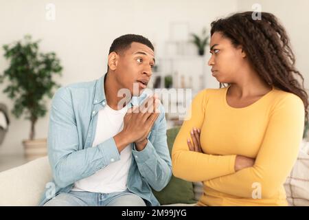 Sad young african american man asking for forgiveness from angry offended female in living room interior Stock Photo