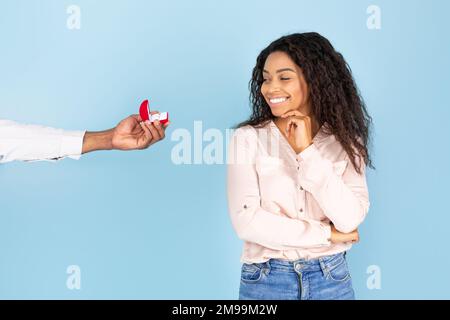 Marriage proposal. Man giving ring in box to happy black lady isolated on blue background, studio shot, copy space Stock Photo