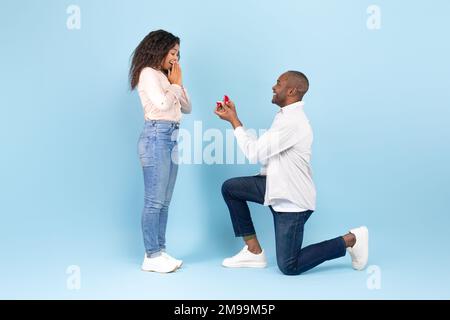 Loving black man standing on one knee and offering engagement ring to his beloved woman on blue background, side view Stock Photo