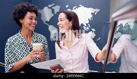 Two Female Colleagues Have Informal Meeting On Stairs Of Modern Office With World Map In Background Stock Photo
