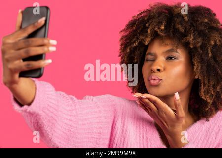 Happy young african american curly woman taking selfie on smartphone and blowing kiss, wink Stock Photo
