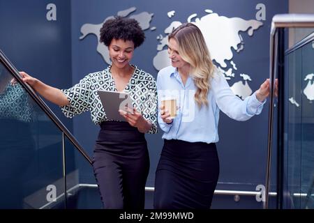 Female Colleagues With Digital Tablet Meeting On Stairs Of Office With World Map In Background Stock Photo