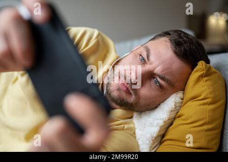 Close-up portrait adult man lying down on sofa at home and playing mobile game using his cell phone. Stock Photo