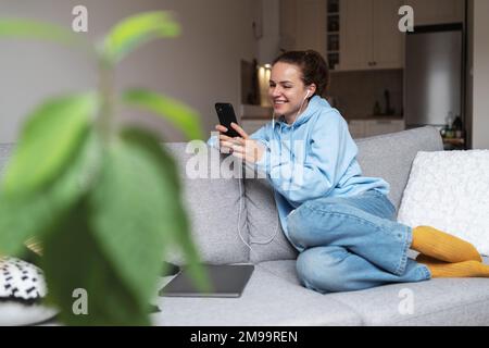 Young brunette woman sitting on sofa at home using smartphone watching video on internet and smiling. Stock Photo