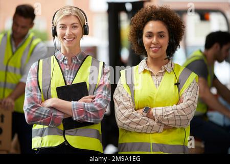 Portrait Of Two Female Workers Using Headsets In Distribution Warehouse With Digital Tablet Stock Photo