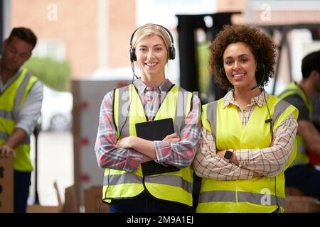 Portrait Of Two Female Workers Using Headsets In Distribution Warehouse With Digital Tablet Stock Photo