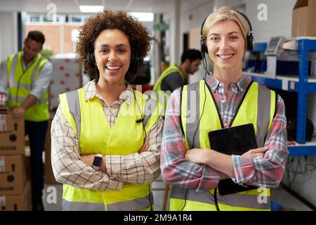 Portrait Of Two Female Workers Using Headsets In Distribution Warehouse With Digital Tablet Stock Photo