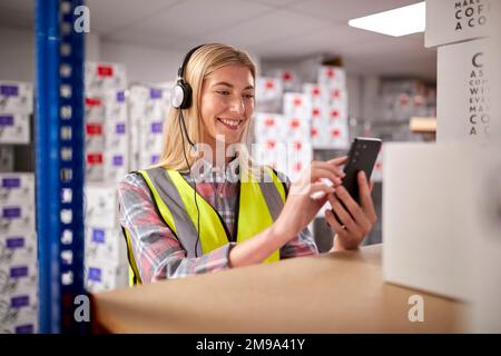 Female Worker Wearing Headset In Logistics Distribution Warehouse Using Mobile Phone Stock Photo
