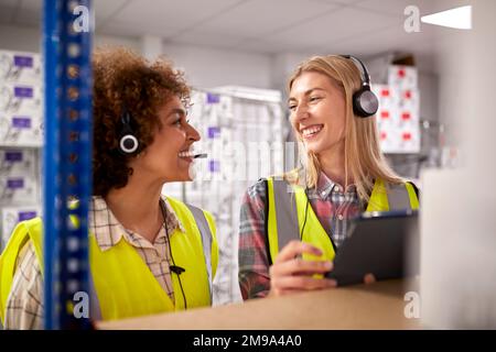 Two Female Workers Wearing Headsets In Logistics Distribution Warehouse Using Digital Tablet Stock Photo