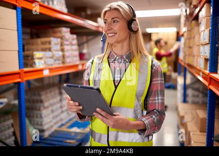 Female Worker Wearing Headset In Logistics Distribution Warehouse Using Digital Tablet Stock Photo