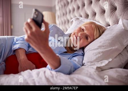 Menopausal Mature Woman Lying In Bed With Hot Water Bottle At Home Using Mobile Phone Stock Photo