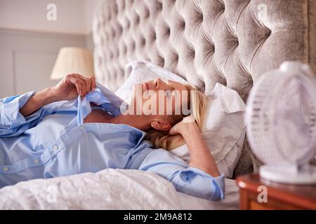 Menopausal Mature Woman Suffering With Insomnia In Bed At Home Using Electric Fan Stock Photo