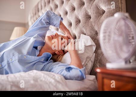 Menopausal Mature Woman Suffering With Insomnia In Bed At Home Using Electric Fan Stock Photo