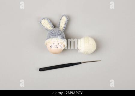 Easter egg with funny face and gray crocheted hat with bunny ears with a balls of thread and a crochet hook on a gray background. Stock Photo