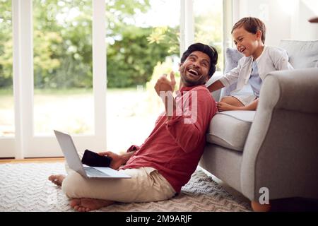 Man Working From Home Sitting On Floor In Lounge Using Laptop And Mobile Phone As Son Watches Stock Photo