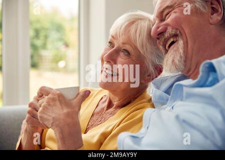 Retired Senior Couple Sitting On Sofa At Home Drinking Coffee And Watching TV Together Stock Photo