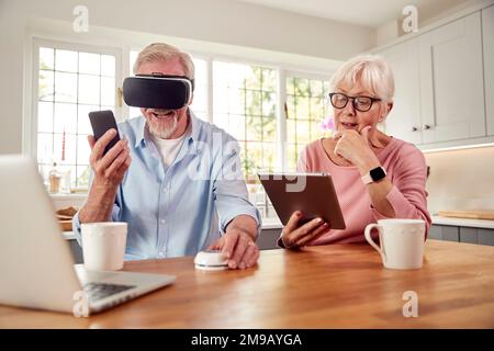 Retired Senior Couple In At Home Using Digital Tablet VR Headset Smartwatch Mobile Phone And Laptop Stock Photo