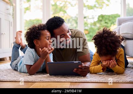 Father At Home Lying On Rug In Lounge With Children Using Digital Tablet Stock Photo