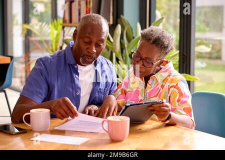 Senior Couple Sitting Around Table At Home Reviewing Finances Using Digital Tablet Stock Photo