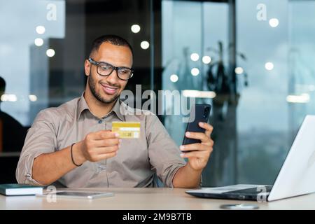 Successful hispanic businessman inside office, man in shirt smiling and happy holding bank credit card and phone, making money transfer and online shopping. Stock Photo