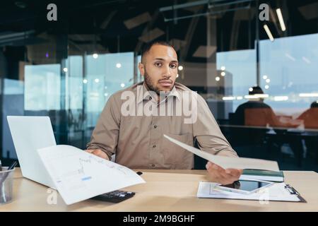 Dissatisfied businessman shows documents financial reports to camera, frustrated financier behind paper work looks at camera upset, latin american man working inside office on laptop. Stock Photo