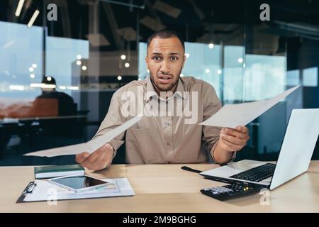 Dissatisfied businessman shows documents financial reports to camera, frustrated financier behind paper work looks at camera upset, latin american man working inside office on laptop. Stock Photo