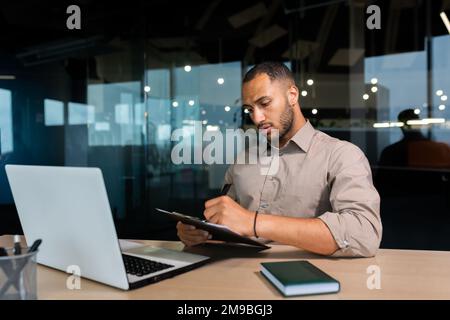 Serious concentrated businessman writing document while sitting in office using laptop for online learning, hispanic man in glasses listening to video call report at workplace. Stock Photo
