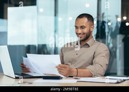Successful businessman inside the office reading a financial report, hispanic man holding a contract in his hands and smiling with satisfaction, good achievement results, working on a laptop. Stock Photo