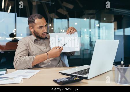 Dissatisfied businessman shows a graph with bad achievements to the camera on a video call, a Latin American man reports negative results at an online meeting of company managers, a man at work. Stock Photo