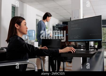 Web developer in wheelchair working on server coding in big data office, analyzing programming language and script on terminal window. Disabled woman programmer with impairment working on it system. Stock Photo