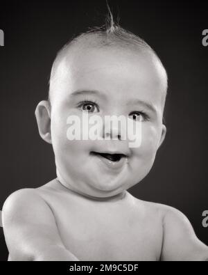 1950s BABY WITH QUIRKY FACIAL EXPRESSION LOOKING AT CAMERA  - b10088 HAR001 HARS B&W EYE CONTACT HUMOROUS HAPPINESS HEAD AND SHOULDERS CHEERFUL COMICAL PRIDE SMILES TWIST COMEDY JOYFUL KOOKY JUVENILES OFFBEAT QUIRKY BABY GIRL BLACK AND WHITE CAUCASIAN ETHNICITY EXPRESSIVE HAR001 OLD FASHIONED Stock Photo