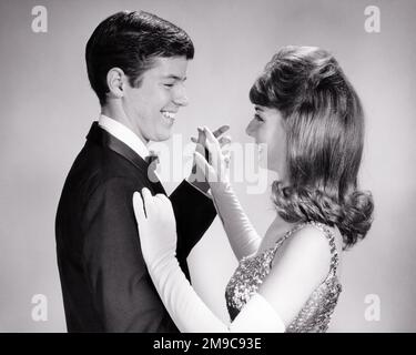 1960s TEEN COUPLE FORMAL ATTIRE DANCING ARM IN ARM AT SCHOOL PROM  - d5536 HAR001 HARS STYLE YOUNG ADULT TEAMWORK PLEASED JOY LIFESTYLE CELEBRATION FEMALES STUDIO SHOT HEALTHINESS COPY SPACE FRIENDSHIP HALF-LENGTH PERSONS MALES TEENAGE GIRL TEENAGE BOY ENTERTAINMENT CONFIDENCE B&W DATING SCHOOLS TUX HAPPINESS CHEERFUL ARM IN ARM HAIRSTYLE ATTRACTION HIGH SCHOOL SMILES HIGH SCHOOLS CONNECTION COURTSHIP JOYFUL STYLISH TEENAGED ATTIRE SEQUINS COOPERATION JUVENILES LONG WHITE GLOVES SOCIAL ACTIVITY TOGETHERNESS YOUNG ADULT MAN YOUNG ADULT WOMAN BIG HAIR BLACK AND WHITE CAUCASIAN ETHNICITY COURTING Stock Photo