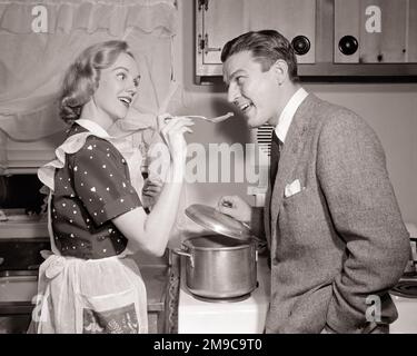 https://l450v.alamy.com/450v/2m9c9t0/1950s-woman-housewife-in-print-dress-and-apron-holding-tasting-spoon-to-man-husbands-mouth-in-kitchen-h2939-deb001-hars-suburban-urban-relationship-old-time-nostalgia-old-fashion-print-1-silly-well-communication-comic-teamwork-testing-pleased-joy-lifestyle-females-married-spouse-husbands-home-life-half-length-ladies-persons-males-bw-partner-homemaker-temptation-suit-and-tie-humorous-happiness-homemakers-cheerful-courage-and-choice-comical-pride-tasting-in-to-authority-housewives-relationships-smiles-who-conceptual-comedy-joyful-lid-support-deb001-mans-cooperation-mid-mid-adult-2m9c9t0.jpg