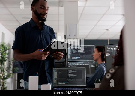 Software developer holding clipboard checking html code on terminal window, using server database on computer. Coders talking about programming language and cloud computing user interface. Stock Photo