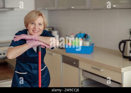 Portrait of smiling senior housekeeping standing with mop on kitchen and looking at camera Stock Photo