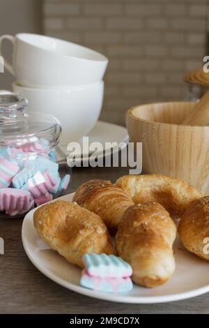 Croissants, marshmallows and tea for teatime on a wooden table, white brick in the background. Stock Photo