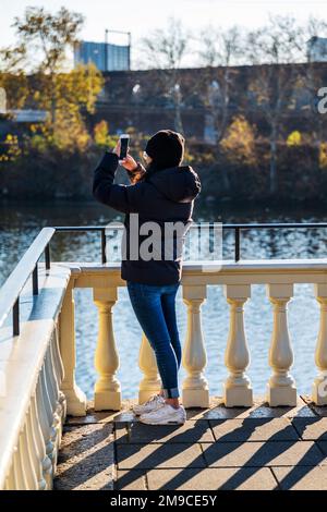 Young woman using iPhone to photograph along sunlit stone path; Fairmount Water Works; Schuylkill River; Philadelphia; Pennsylvania; USA Stock Photo