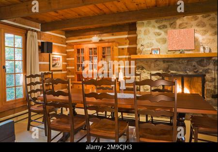 Long wooden dining table with high back chairs next to lit fieldstone fireplace in dining room inside Canadiana style log home. Stock Photo