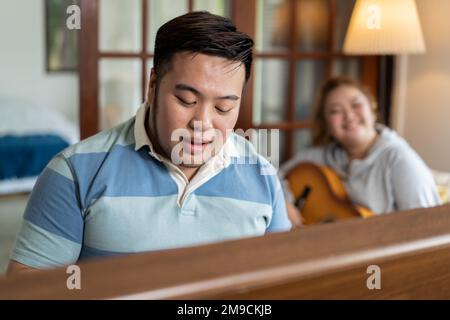 Young Asian chubby couple singing and playing acoustic guitar and piano together. Man and woman enjoying musical instrument. People in a band practicing in the house Stock Photo