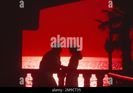 A couple silhouetted standing on the balcony next to the railing with a red-hot sunset behind them in the ocean Stock Photo