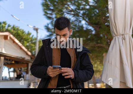 Serious bearded guy in outerwear browsing smartphone while chatting on social media during free time in outdoor cafeteria Stock Photo