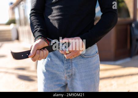 Crop anonymous male in black turtleneck and jeans standing on street in sunny weather while fastening belt on trousers Stock Photo