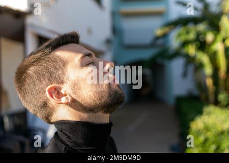 Side view of calm bearded guy with earring and closed eyes leaning head back while standing against blurred background of street in sunny weather Stock Photo
