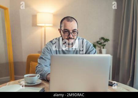 Portrait of young bearded man in trendy spectacles working at laptop at home office. Stock Photo