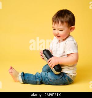 Toddler baby plays with a wireless music speaker on a studio yellow background. Happy child in a white t-shirt and blue jeans listens to music in an a Stock Photo