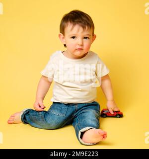 Toddler baby plays with toy cars on a studio yellow background. Happy child in white t-shirt and blue jeans plays with toy cars. Kid aged one year fou Stock Photo