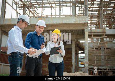 Asian Architector and engineers team with draft plan of building and tablet talking constructing site. Construction manager and engineer working on bu Stock Photo
